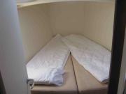 Ein V-förmiges Bett mit zwei beigen Matrazen worauf zwei weiße Decken liegen. Das Bett geht bis an die Bordwand, die mit einem beigen Stoff verkleidet ist.
