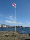 Auf einer Felsfläche nahe dem Wasser und nur wenig höher als der Wasserspiegel weht an einem weißen Flaggenmast die Flagge der Faröer. Im Hintergrund auf der anderen Seite des Hafenbeckens sind Gebäude zu sehen.