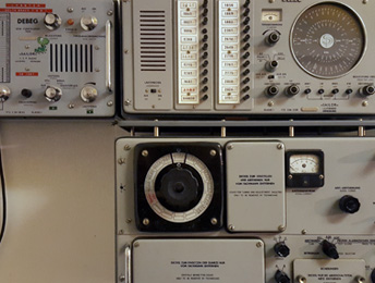Drei graufarbene Funkgeräte an einer Wand sind zu erkennen, ausgestattet mit zahlreichen Schaltern, Drehknöpfen und analogen Rundanzeigen. Mit diesen Funkgeräten kann man über Ozeane hinweg um die ganze Welt funken.