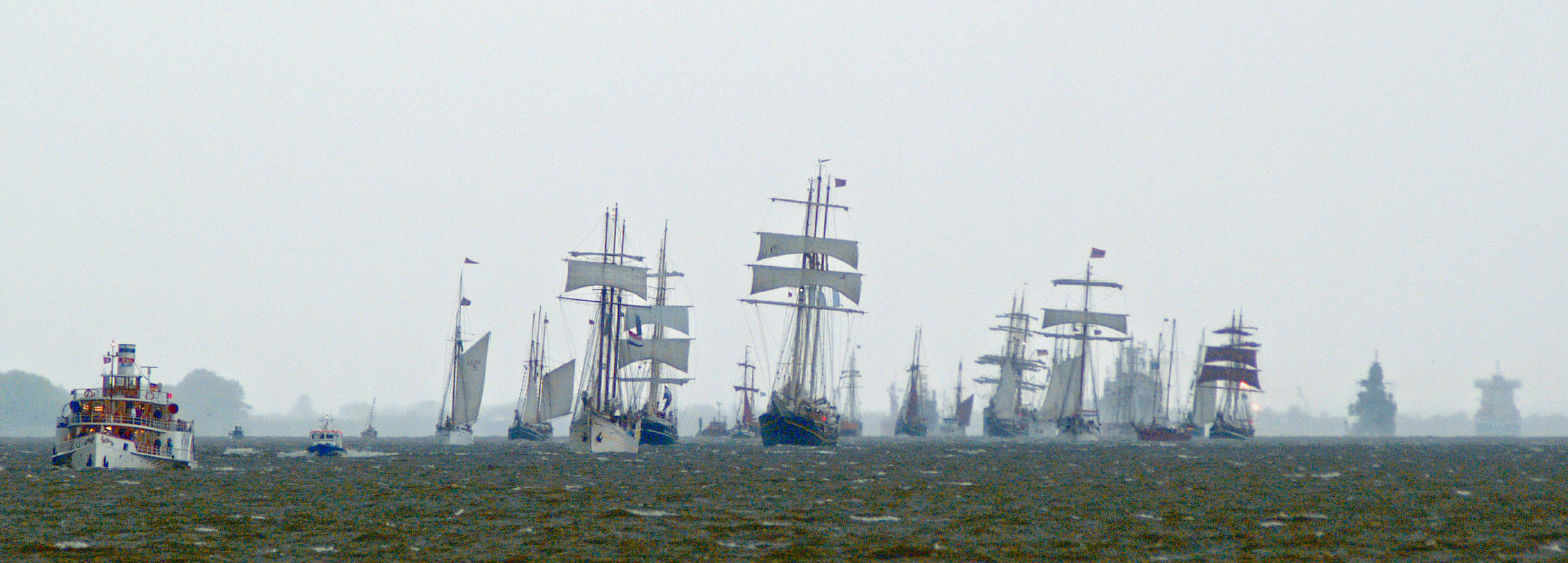 Auf breiter Front segeln historische Rahsegler auf der Elbe auf den Betrachter zu. Im Vordergrund ist eine historische und reich verzierte Dampfbarkasse zu sehen. Alle Segelschiffe krängen zum rechten Bildrand, da eine Bö über die Szene weht.