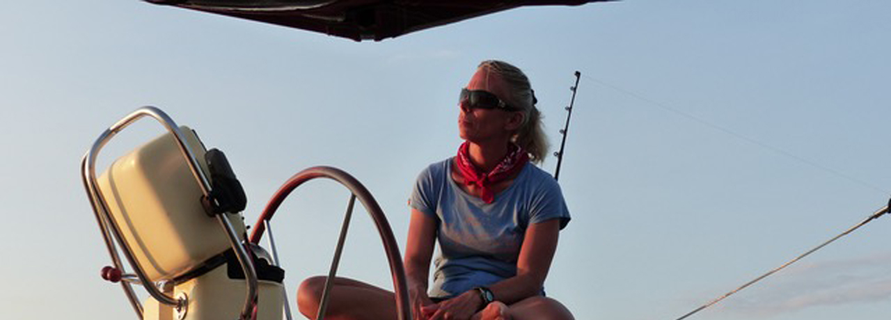 Die blonde Mareike Guhr sitzt in Sommerkleidung lässig mit dem angewinkelten einen Bein, über das sie das andere Bein geschlagen hat, auf dem Steuersitz ihres Katamarans. Vor ihr die Steuersäule mit Ruderrad und Seekartenplotter. Im Hintergrund das Meer.