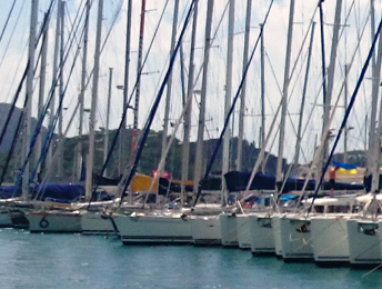 Eine große Anzahl Einrumpf- und Mehrrumpfsegelyachten liegen mit dem Heck zu Steg nebeneinander in einem Yachthafen. Die meisten sind weiß und haben Aluminiummasten. Im Hintergrund sind drei bewaldete Berggipfel zu sehen.