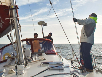 Eine moderne Segelyacht auf dem Meer von vorne nach achtern fotografiert. Die Sonne lässt das Wasser glitzern. Am Want in der Bootsmitte steht ein Mensch und schaut in Mast hinauf. Im Cockpit hinter der Sprayhood gucken zwei Leute nach vorne.