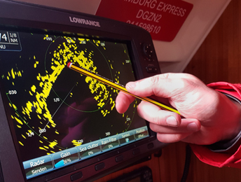 Ein Radarbildschirm mit der Darstellung der Radarechos in Gelb vor schwarzem Hintergrund. Eine Hand an einem mit einer Segeljacke bekleideten Arm zeigt mit einem Bleistift auf eine Stelle auf dem Bildschirm.